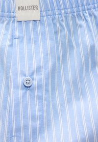 Ανοιχτό μπλε ύφασμα με κάθετες λευκές ρίγες, ένα μπλε κουμπί και μια λευκή ετικέτα μάρκας Hollister ραμμένη κοντά στη μέση.