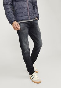 Temno modri kavbojke s krojem slim-fit, ki imajo zbledela mesta in ravne hlačnice. Nosijo se z temnomodrim napihljivim jopičem in belimi športnimi čevlji.