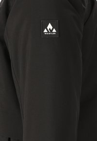 Sorte bukser med en glat tekstur og et patch på låret, der viser "Whistler"-logoet i hvid, fremhævet af geometriske former.