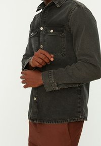 Camicia di denim nero con due tasche frontali, bottoni a scatto e colletto, caratterizzata da una texture leggermente sbiadita e una vestibilità morbida.