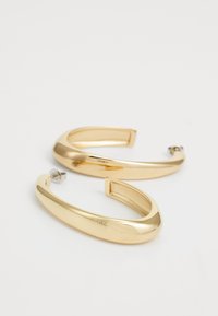 Orecchini a cerchio color oro realizzati in metallo liscio con una forma arrotondata e un design aperto. Presentano un logo discreto inciso su un lato.