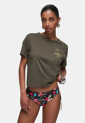 Donna che indossa una t-shirt corta verde oliva con la scritta "golden hour" e slip bikini neri con stampa floreale, accessoriata con orecchini e bracciale argentati.