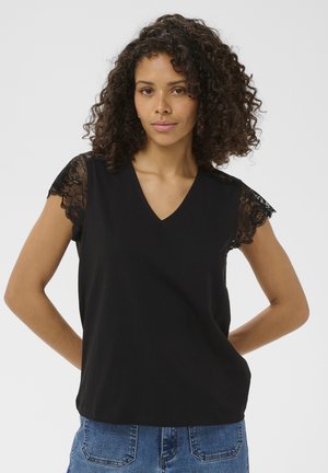 Femeie cu păr creț, purtând un top negru fără mâneci cu umeri din dantelă și blugi albaștri, stând cu mâinile la spate.