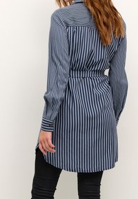 Camicetta a righe blu navy e azzurro con maniche lunghe, colletto e cintura legata in vita. Il tessuto sembra liscio con una vestibilità strutturata.