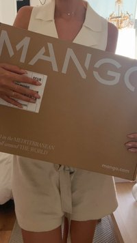 Hnědá kartonová krabice s bílým logem "MANGO", která obsahuje přepravní štítek. Světlé oblečení s uvázaným páskem. Vnitřní prostředí.