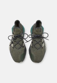 Pár olivově zelených pletených tenisek s tmavě šedými tkaničkami v mašlích, bílými bočními pruhy a texturovanými černými podrážkami na bílém pozadí.