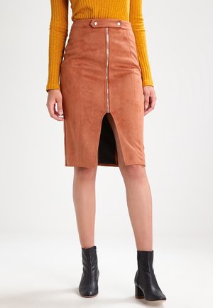 Kobieta ubrana w brązową zamszową ołówkową spódnicę z przednim zamkiem błyskawicznym i rozcięciem, musztardowy prążkowany top z długim rękawem oraz czarne botki za kostkę.