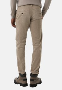 Beżowe spodnie z korduroy o prostym kroju, z tylnymi kieszeniami zapinanymi na czarne guziki, noszone z szaro-czarnymi butami turystycznymi.