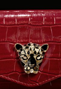 Czerwona torebka z sztucznej skóry krokodyla z zapięciem w kształcie jaguara w złotym kolorze, ozdobionym czarnymi i przezroczystymi kryształkami. Gładka, błyszcząca tekstura.