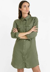 Robe chemise vert olive avec des boutons, des poches plaquées et un col. Fabriquée en tissu doux, avec des manches roulées et un ourlet courbé.