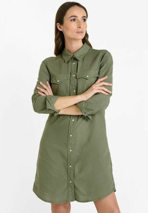Olivgrünes Hemdkleid mit Knöpfen, aufgesetzten Taschen und einem kragenlosen Ausschnitt. Aus weichem Stoff gefertigt, mit hochgekrempelten Ärmeln und einem geschwungenen Saum.