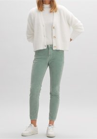 Cardigan blanco de felpa con cuatro botones, combinado con pantalones de pana verde claro. El modelo lleva zapatillas blancas y un suéter de cuello alto blanco ajustado.