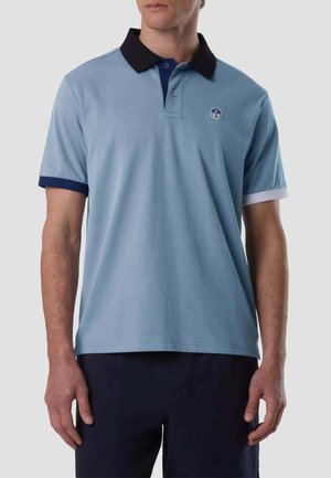 Uomo che indossa una polo azzurro chiaro con colletto nero, polsini blu navy e bianchi, e piccolo logo rotondo sul petto sinistro.