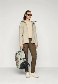 Μπεζ αδιάβροχο μπουφάν με κουκούλα, συνδυασμένο με κρέμ πουλόβερ και καφέ παντελόνια. Γκρι και μαύρο σακίδιο με πολλές θήκες, φορεμένο με casual στυλ.