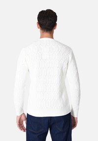 Maglione bianco lavorato a maglia con un motivo a diamante, scollatura e polsini a coste, con dettagli testurizzati visibili sul retro. Abbinato a jeans scuri.