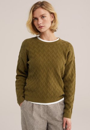 Femme blonde portant un pull en maille texturée vert olive sur un haut blanc et un pantalon gris plissé, debout devant un fond neutre.