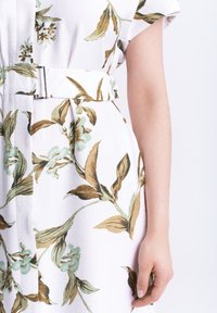 Sezione di un vestito bianco con stampa floreale verde e marrone e cintura coordinata, che mostra un braccio nudo e la mano rilassata al lato.