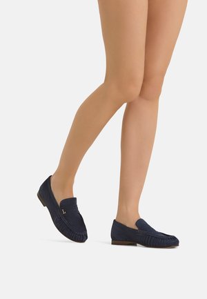 Legs wearing sheer nude tights and navy blue suede loafers with low heels on a white background.