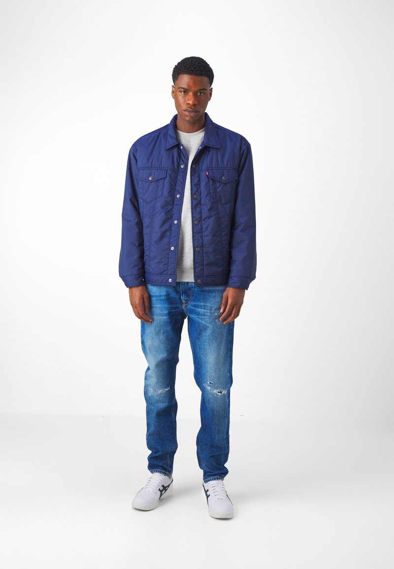 Chaqueta de quilt en color navy con dos bolsillos en el pecho, camiseta gris debajo, combinada con jeans claros desgastados y zapatillas blancas.