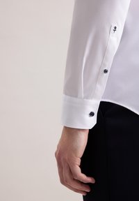 Camicia bianca con texture liscia, dotata di polsini abbottonati con bottoni scuri a contrasto e delicato ricamo floreale sulla manica.