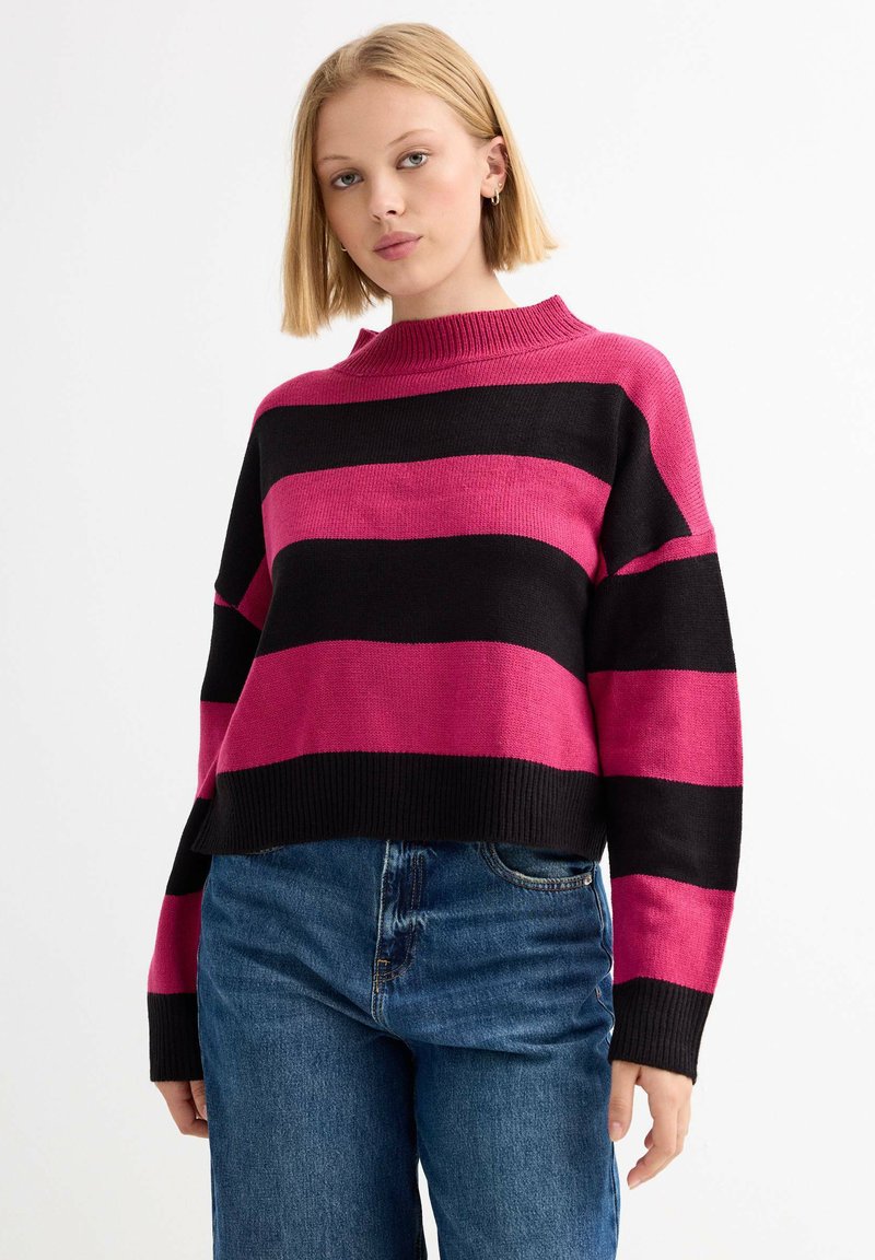 Suéter de punto recortado con amplias rayas negras y rosas, cuello acanalado alto y mangas sueltas. Combinado con jeans de tiro alto azules.