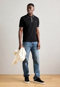 Polo noir avec col et accents de manche blancs, associé à un jean bleu clair et des baskets noires, tenant un pull blanc.