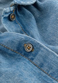 Helles blaues Jeansmaterial mit einem Knopf im Vierlochdesign und sichtbaren Nähten. Die Textur ist weich und hat einen dezenten Glanz.