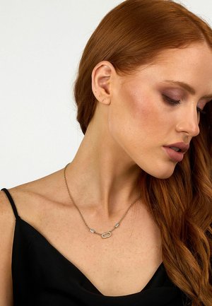 Collana in oro con maglie rettangolari e ovali decorate con dettagli trasparenti. La modella indossa un top nero, evidenziando il design della collana.