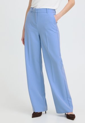 Pantaloni a gamba larga azzurri chiaro indossati con un top bianco, mano del modello in tasca, abbinati a décolleté marroni con punta affilata.