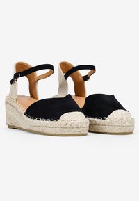 Sandalias de esparto de ante negro con tiras de tela trenzada, plataforma de cuerda natural y parte trasera abierta. La suela de goma proporciona tracción y estabilidad.