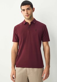 Burgundy színű, rövid ujjú polo póló, gallérral és három gombos kivágással. A bal mellrészen egy kis fekete logó található. Pamut keverék anyagból készült.