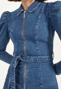 Chaqueta de denim con cierre de cremallera, mangas abullonadas y un cinturón atado en la cintura. La tela es de un azul medio y tiene una textura suave.