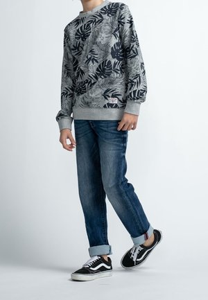 Enfant portant un sweat-shirt gris avec des motifs de feuilles noirs, un jean bleu retroussé et des baskets noir et blanc, debout sur un fond uni.