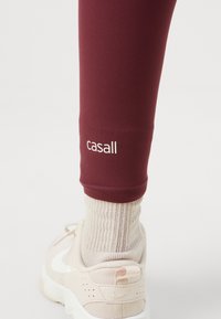 Vinröda träningsleggings med logotypdetalj vid fållen, matchade med ljusbeiga sportskor och krämfärgade ribbade strumpor. Slät och elastisk material.