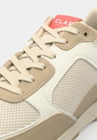 Beige athletic shoe featuring a mesh upper, smooth leather accents, and a red label. Lace-up design with textured patterns and stitching details.