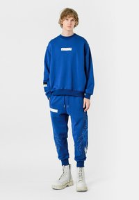 Conjunto de sudadera y pantalones deportivos azul, con una tela texturizada, puños acanalados y detalles gráficos en los pantalones. Combinado con botas de combate blancas.