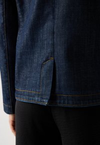 Nærbilde av kanten på en mørkeblå denimjakke med oransje sømmer over svarte bukser, som viser sideslitt detalj og en del av venstre arm.