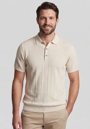 Man with short brown hair and beard wearing a beige short-sleeve polo shirt and light brown pants, standing with one hand in pocket.