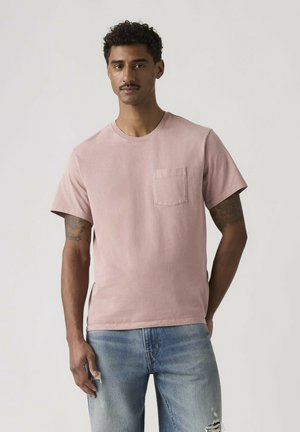 Man with short curly hair and tattoos wearing a light pink pocketed T-shirt and distressed blue jeans, standing with one hand behind back.