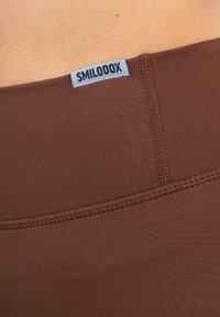 Bruine elastische leggings met een gladde textuur, voorzien van een gestikte naad en een klein blauw label met "SMILODOX" op de tailleband.
