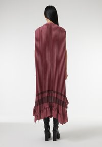 Robe plissée longue en prune avec un ourlet à volants et des accents transparents. Texture lisse, manches courtes, tombe jusqu'aux chevilles. Portée avec des bottes noires imposantes.