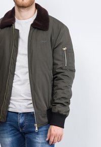 Olivgrüner Bomberjacke mit einem braunen Kunstfellkragen, Reißverschlusstaschen, Rippbündchen und einer gesteppten Fütterung. Verfügt über eine glatte Nylon-Oberfläche.