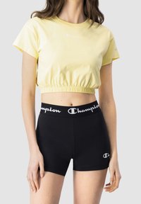 Žena nosí žluté zkrácené tričko a černé kraťasy s vysokým pasem, na kterých je logo "Champion" na pásu a nohavici.
