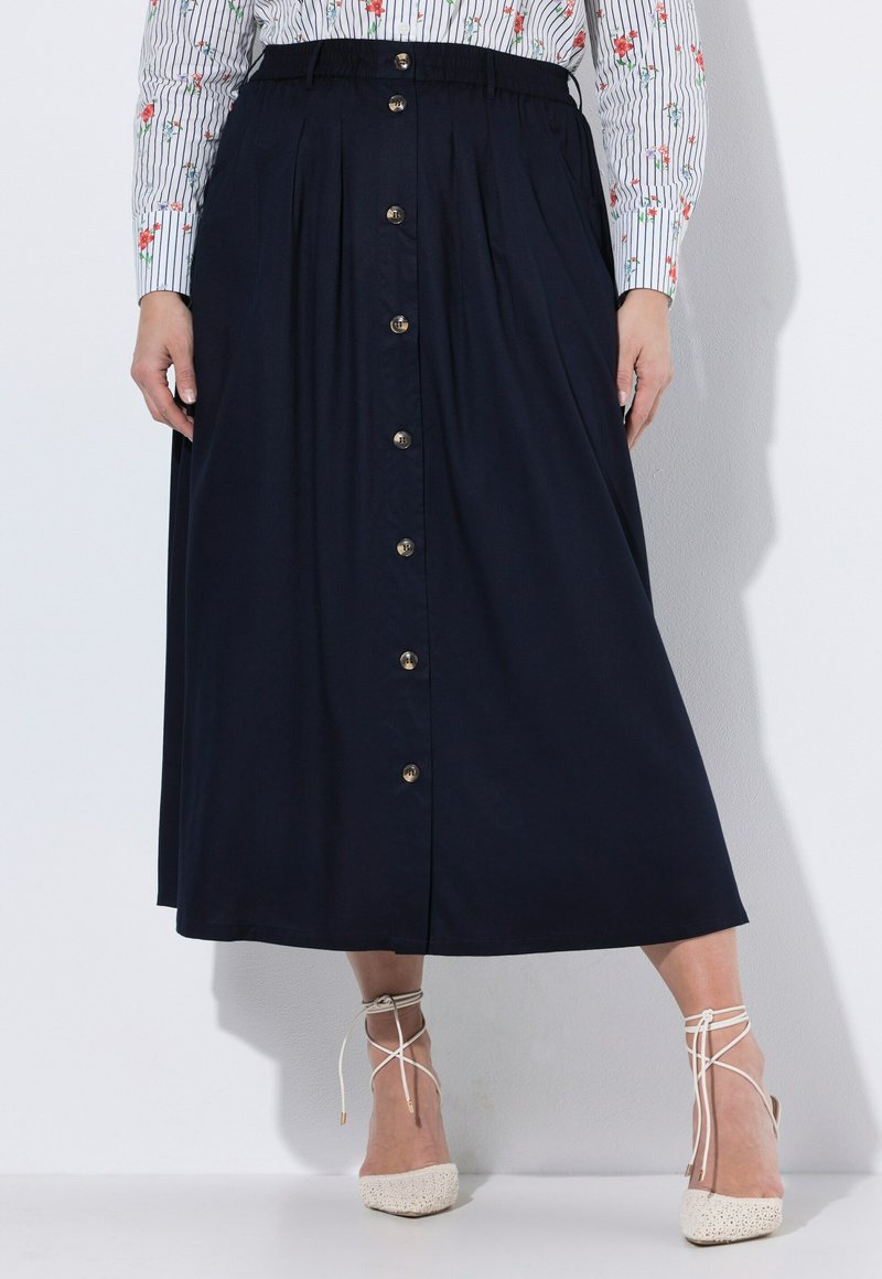 Frau trägt einen midilangen marineblauen Rock mit vorderen Knöpfen, kombiniert mit einem gestreiften Blumenhemd und weißen Schnürabsätzen.