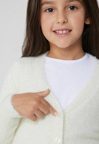 Hellgrüner, flauschiger Cardigan mit V-Ausschnitt und langen Ärmeln, der vorne einen klaren Knopf hat. Das Modell zeigt auf den Knopf.