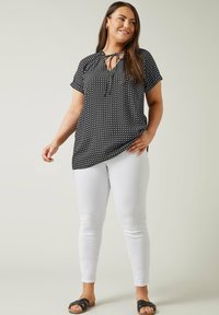 Schwarze Bluse mit einem Punkte-Muster, kurzen Ärmeln und einem Schlitz-Krawatten-Ausschnitt. Kombiniert mit weißen Skinny-Jeans und schwarzen Sandalen.