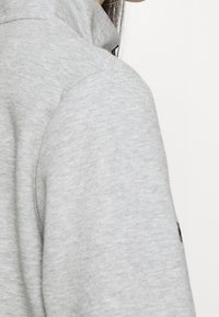 Ljusgrå sweatshirt med en mjuk textur, som har en hög krage och dragkedjedetalj. Tyget ser mjukt och avslappnat ut.