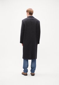 Dunkler Wollmantel mit strukturiertem Finish, langen Ärmeln, Reverskragen, einem hinteren Schlitz, getragen über blauen Jeans und braunen Knöchelstiefeln.