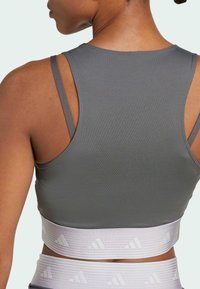 Gray fitted athletic sports bra with a wide light gray band featuring a subtle logo pattern and adjustable straps. Smooth texture.