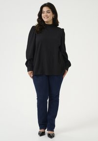 Zwarte blouse van textuur met pofmouwen en hoge halslijn, gecombineerd met donkerblauwe strakke jeans en zwarte puntige schoenen.